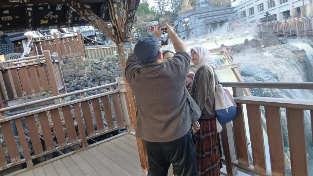 Suasana ramai dan penuh keceriaan di Yubatake Kusatsu Onsen, wisatawan berjalan santai, berfoto, dan menikmati uap air panas di tengah area onsen yang hidup dan hangat